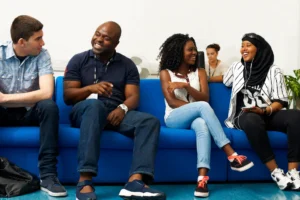 Four people chat and laugh on a blue sofa at a Leeds community centre, with another working in the background. Doing Good Leeds.