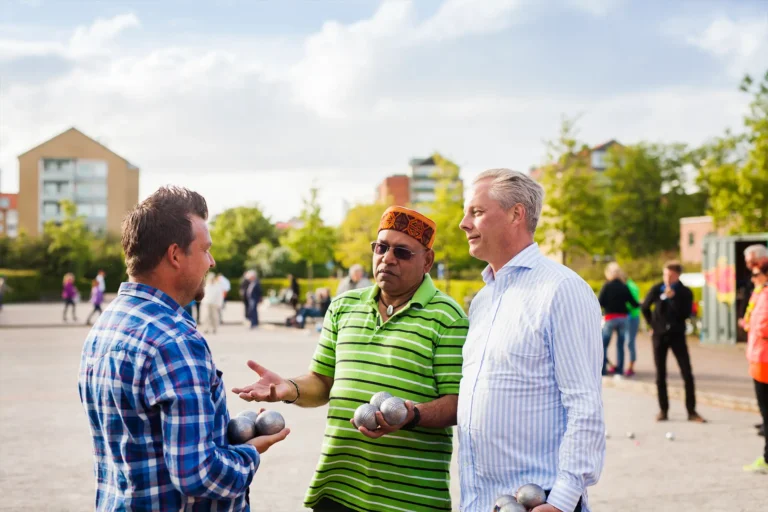 Three men chat while holding pétanque balls outdoors in Leeds; community event with people and buildings in the background.