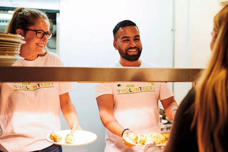 Two smiling volunteers serve food from behind a counter to a person at a Leeds charity kitchen. Plates in hand, they help their community.
